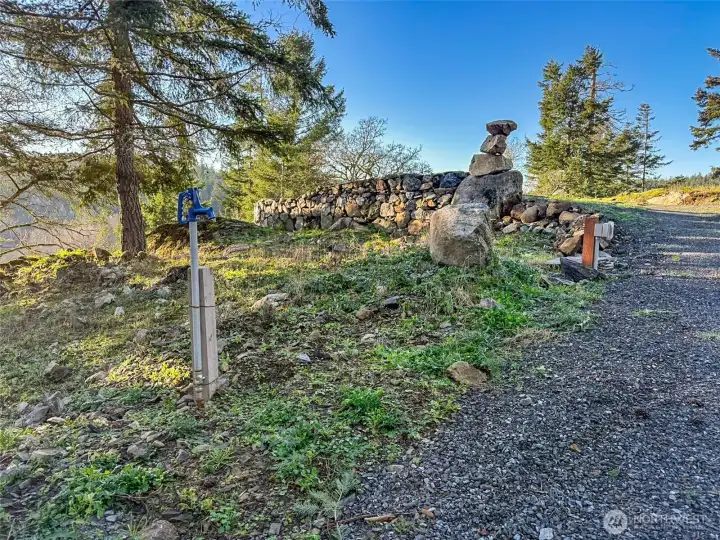 Approach to the building site and upper patio via a gravel access drive, featuring an electrical service pedestal, frost-free yard hydrant, handcrafted stone walls, and mature trees.