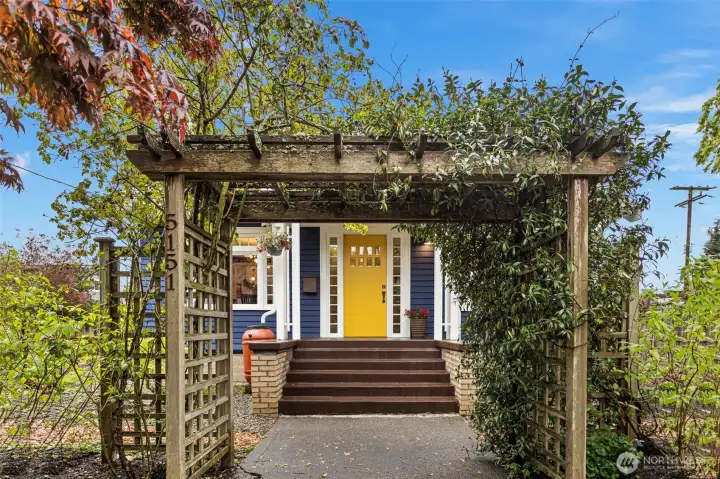5151 on the post, jasmine overhead. The arbor frames the entry — and the yellow door earns the walk.