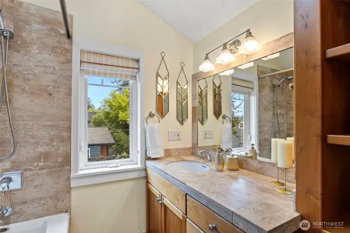 Travertine tile, wood vanity, jetted tub, handheld shower, skylight overhead, built-in linen shelving. The primary bath doesn't ask for much — just blue sky and hot water.