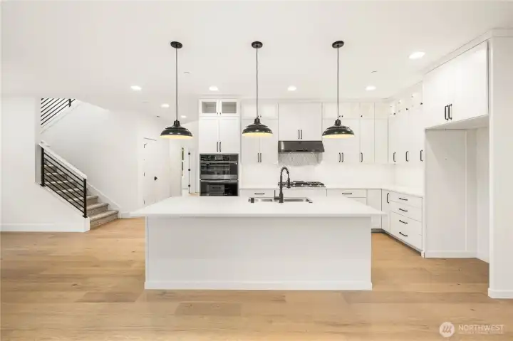 This view showcases the full kitchen layout along with the open railing staircase that enhances the home’s airy, modern feel. The oversized island and extensive cabinetry offer both style and storage, while the open flow supports seamless movement between the main living spaces. It’s a bright, spacious hub designed for modern Eastside living.  These photos are from Lot 3 at Gooden Hollow, which is the same floorplan. However, Lot 6 will have changes, so all photos, renderings, site maps, and floorplans are for visualization purposes only. Plans, colors, building orientation, finishes, home outlook, and other details can and WILL vary.