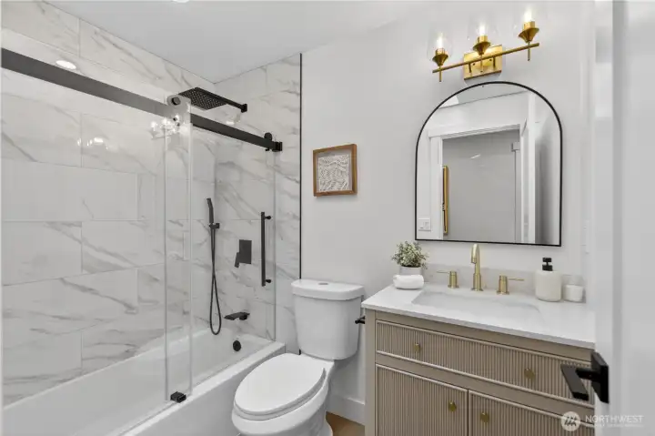 Bright white walls and a skylight bring natural light into this bath, creating a space that feels bright, elevated, and beautifully refreshed. Glass shower doors showcase marbled shower tile that echoes the kitchen finishes, giving the home a cohesive designer feel, while black shower hardware, a rainfall shower head, and handheld sprayer add modern comfort. A fluted furniture-style vanity brings texture and generous storage, topped with white quartz that reflects the kitchen’s clean, refined surfaces. Gold-tone swan-neck faucets, an arched black-framed mirror, and elegant glass-shade vanity lighting complete the room with warmth and polish. Bright white walls and a skylight bring natural light into this bath, creating a space that feels bright, elevated, and beautifully refreshed. Glass shower doors showcase marbled shower tile that echoes the kitchen finishes, giving the home a cohesive designer feel, while black shower hardware, a rainfall shower head, and handheld sprayer add modern comfort. A fluted furniture-style vanity brings texture and generous storage, topped with white quartz that reflects the kitchen’s clean, refined surfaces. Gold-tone swan-neck faucets, an arched black-framed mirror, and elegant glass-shade vanity lighting complete the room with warmth and polish.