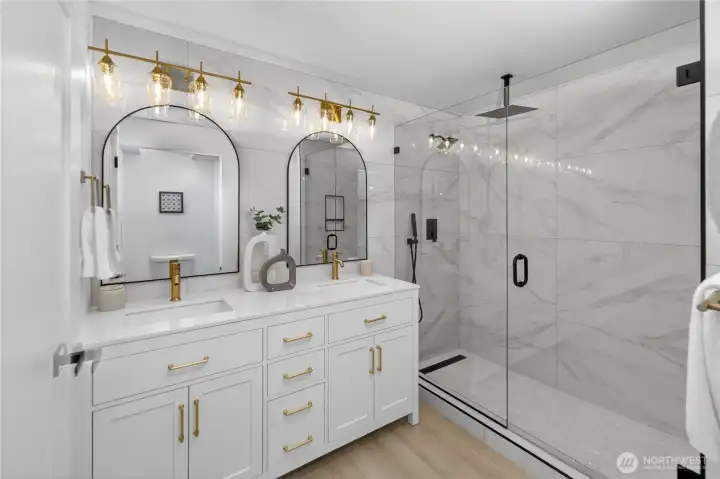 Bright white walls and a skylight bring natural light into the primary bath, creating an uplifting, spa-like feel. Floor-to-ceiling marbled tile wraps the large walk-in shower in beauty, with glass walls, black-tone fixtures, dual rainfall shower heads, and a handheld sprayer for a luxurious everyday experience. The double vanity reflects the kitchen and guest bath finishes with a white quartz counter, crisp cabinet style, generous storage, and spacious double sinks. Champagne-tone vintage-inspired faucets coordinate beautifully with the elegant glass-shade vanity lights, while arched black-framed mirrors complete the room with a refined designer finish. Bright white walls and a skylight bring natural light into the primary bath, creating an uplifting, spa-like feel. Floor-to-ceiling marbled tile wraps the large walk-in shower in beauty, with glass walls, black-tone fixtures, dual rainfall shower heads, and a handheld sprayer for a luxurious everyday experience. The double vanity reflects the kitchen and guest bath finishes with a white quartz counter, crisp cabinet style, generous storage, and spacious double sinks. Champagne-tone vintage-inspired faucets coordinate beautifully with the elegant glass-shade vanity lights, while arched black-framed mirrors complete the room with a refined designer finish.