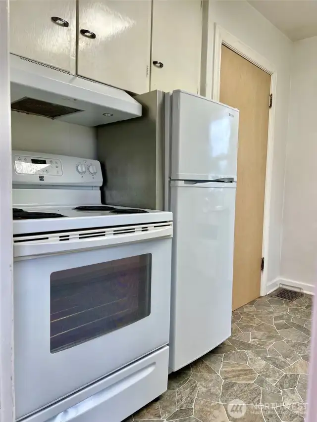 View from the entry of the kitchen showing the range, refrigerator & cabinetry.  The door shown is the door to the downstairs/basement & garage