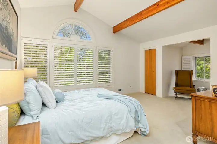 The primary bedroom is on the upper level, it is spacious and features custom windows, vaulted ceiling and wood beams. Note the small sitting area as a cozy nook.