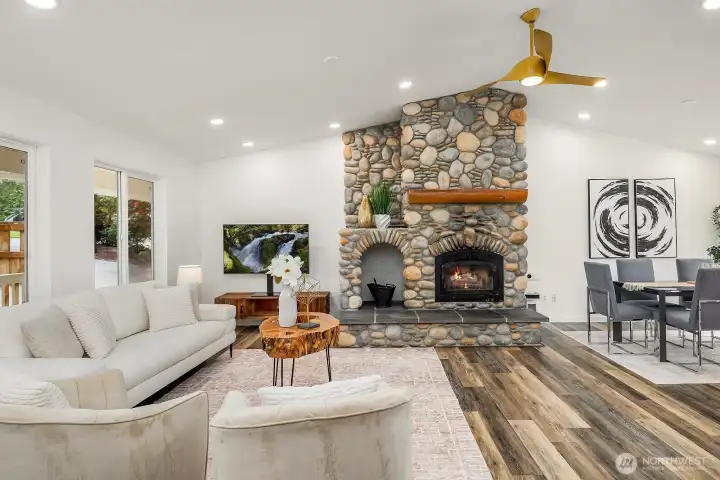 This view of the living room captures the full grandeur of the vaulted ceilings and stone fireplace alongside the home's thoughtful updates, with natural light pouring in from multiple directions to create a warm and welcoming atmosphere.