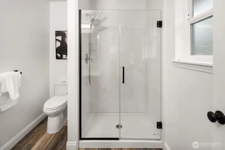 The updated bathroom features a sleek frameless glass shower with matte black hardware, crisp white finishes, and a bright window, delivering a clean, modern aesthetic that complements the home's fresh interior updates throughout.