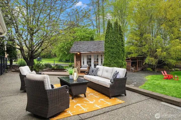 The expansive patio is a true outdoor living room, with a comfortable lounge set surrounded by mature trees, lush landscaping, and the charming outbuilding office just beyond, creating a private and peaceful backyard retreat.