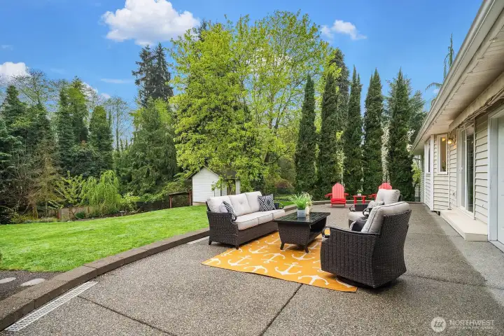 A wider view of the patio showcases its generous size and the stunning mature privacy landscaping that surrounds it, with towering evergreens and lush greenery creating a secluded, resort-like atmosphere perfect for relaxing or entertaining.
