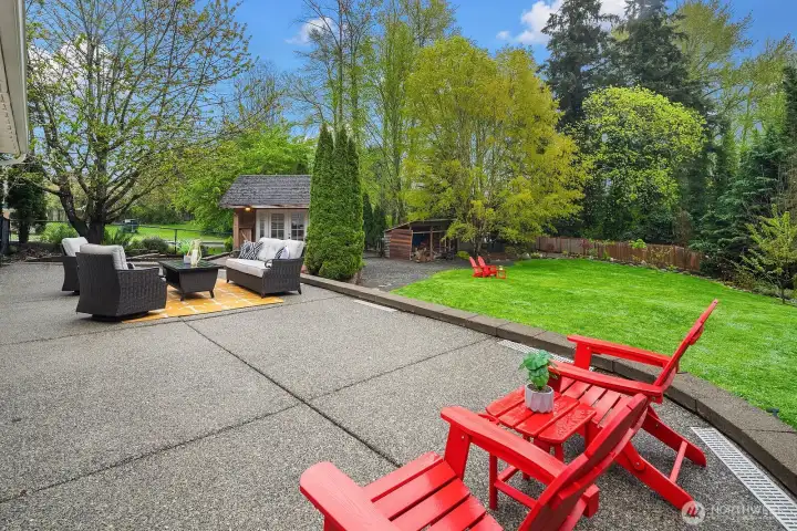 The expansive patio stretches out toward the lush lawn and mature orchard beyond, with cheerful red Adirondack chairs and the charming outbuilding office framed by the property's beautiful mix of evergreens and flowering trees.