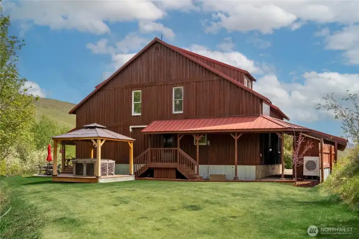 Cedar-sided home with metal roof, carport, and covered deck, complemented by a stone patio, private hot tub, and gazebo—designed for relaxed indoor-outdoor living in a peaceful setting.