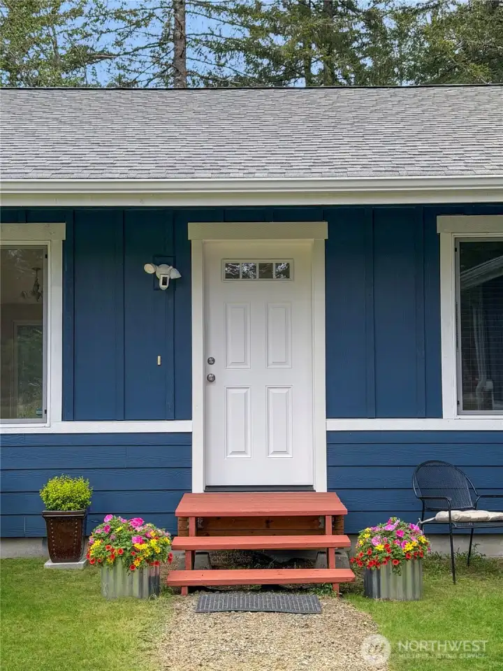 The front porch feels cheerful and welcoming, with a crisp blue exterior, white trim, flowers, and a simple country charm that says, “Come on in.”
