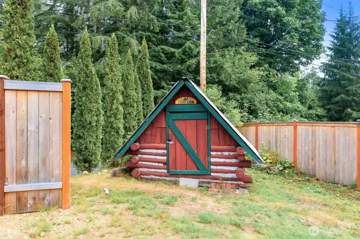 A whimsical, child-sized log cabin playhouse called “Kash’s Kabin,” complete with red logs, green trim, and an A-frame roof—adding personality and charm to the property.