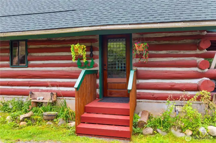 Charming log cabin entry with a solid wood door, colorful hanging baskets, and rustic red-and-white tamarack logs, offering a warm welcome into the home.