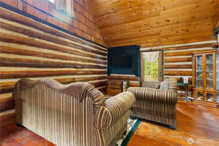 Spacious living room with exposed log walls and vaulted wood-paneled ceilings. A cozy sitting area is centered around the TV and antique-style furnishings, blending rustic comfort with cabin character.
