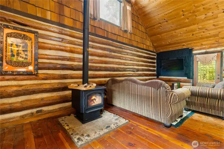 The living room showcases soaring pine ceilings and classic log walls, anchored by a cozy wood-burning stove set on a stone hearth. It’s the perfect place to curl up on a cool evening, with natural light streaming in and enough space for comfortable seating.