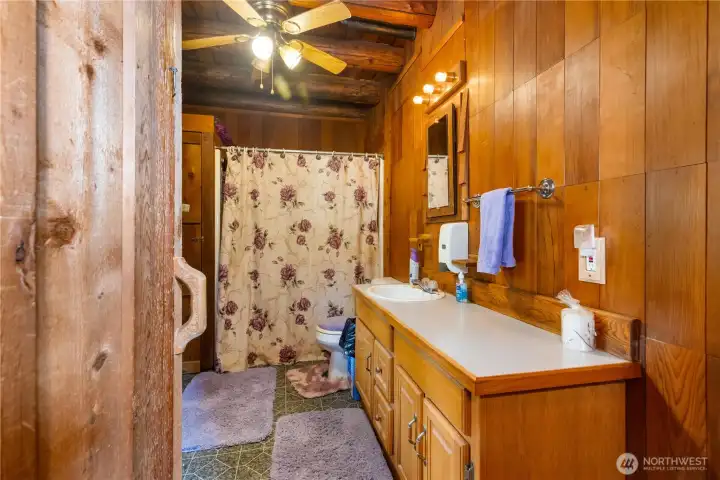 Step into a full bath wrapped in classic wood paneling and log detail, bringing the cabin’s rustic feel into a space that’s both warm and practical. The large vanity offers plenty of storage and counter space, while the floral shower curtain and cozy rugs add a welcoming, homey touch.