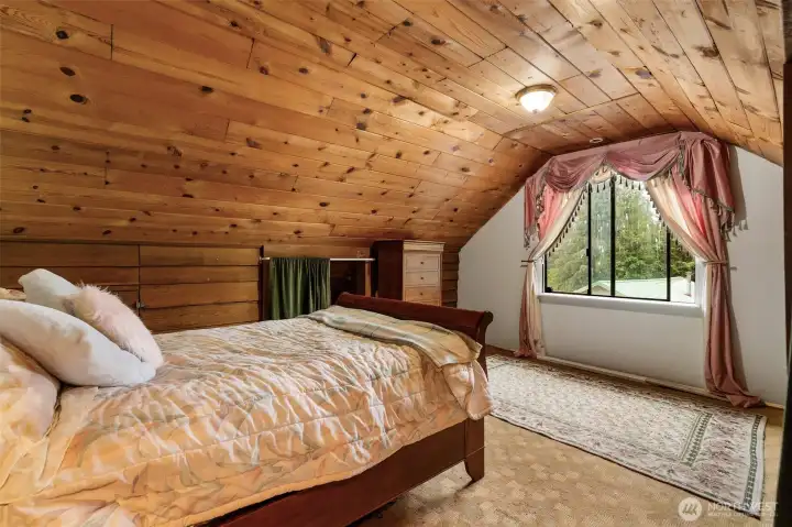 Spacious loft bedroom with sloped wood-paneled ceilings, large window overlooking the trees, and plenty of natural light. A serene retreat with classic cabin character.