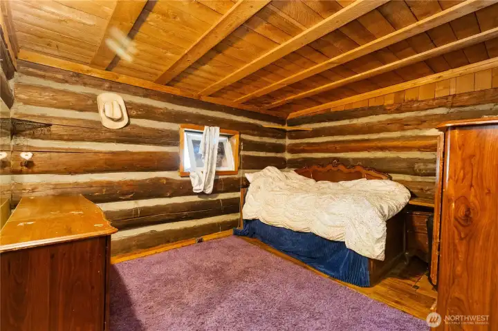 Cozy main-level bedroom with log walls, wood ceiling, and a small window bringing in natural light. Warm and rustic, perfect for a guest room or home office.