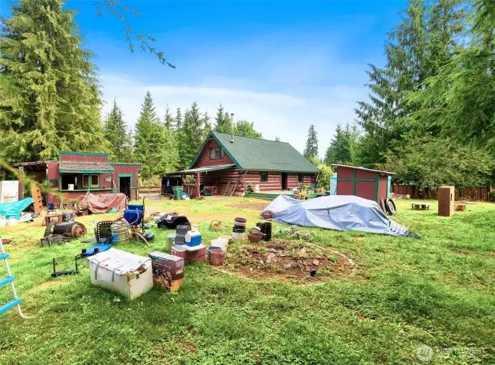 Backyard perspective highlighting the two-story log cabin, rustic saloon-style outbuilding marked “Mercantile,” and multiple sheds. The open yard provides ample space for gardening, projects, and creative outdoor living.