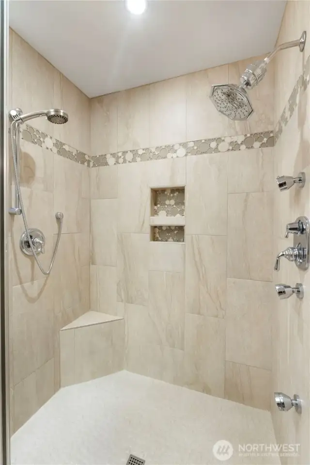 Step into a custom walk-in shower designed for luxury and comfort, featuring dual showerheads, body jets, a built-in corner bench, and sleek tile accents with mosaic inlay. Dual niches provide practical storage, while the frameless glass enclosure adds a modern, open feel.