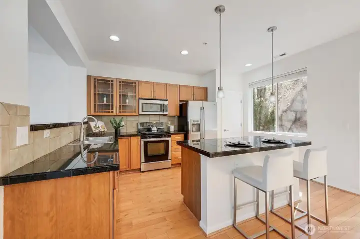 This inviting kitchen features warm wood cabinetry, sleek stainless-steel appliances, and rich granite countertops that offer both style and durability. A spacious center island with bar seating anchors the room, illuminated by modern pendant lighting for an elevated touch. The open layout flows seamlessly into the dining area, enhanced by natural light from the nearby window and complemented by clean, contemporary finishes throughout. It’s a functional and welcoming space designed for everyday living and effortless entertaining.