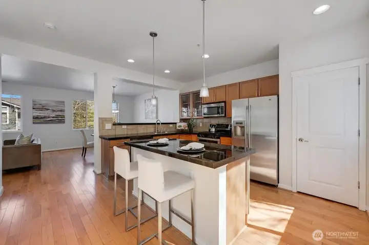 Inviting kitchen features warm wood cabinetry, sleek stainless-steel appliances, and rich granite countertops that offer both style and durability. A spacious center island with bar seating anchors the room, illuminated by modern pendant lighting for an elevated touch. The open layout flows seamlessly into the dining area, enhanced by natural light from the nearby window and complemented by clean, contemporary finishes throughout. It’s a functional and welcoming space designed for everyday living and effortless entertaining.