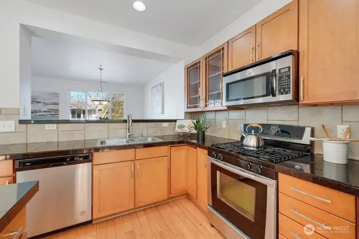 Well-appointed kitchen features light wood cabinetry, sleek stainless-steel appliances, and striking black granite countertops that add both contrast and durability. A neutral tile backsplash ties the space together, while the open pass-through to the adjoining living area brings in natural light and creates an easy, connected flow. Thoughtful details and a clean, contemporary layout make this kitchen both functional and inviting for everyday cooking and entertaining.