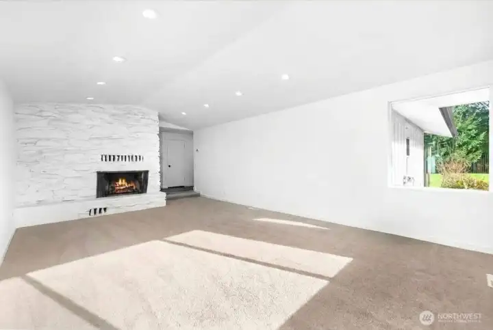 An alternate view of your bright and airy living room with vaulted ceilings, recessed lighting, a cozy wood burning fireplace. Tons of natural light with abundance of windows and space. Hallway past fireplace leads to bedrooms on the right and dining room and kitchen to the left.