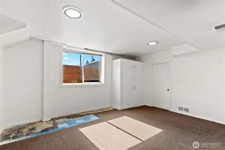 Bedroom with generous natural light from a large window, dark carpeting, and clean white walls. Includes a built-in cabinet for clothing and a discreet door housing the furnace and water tank, making it both functional and comfortable.