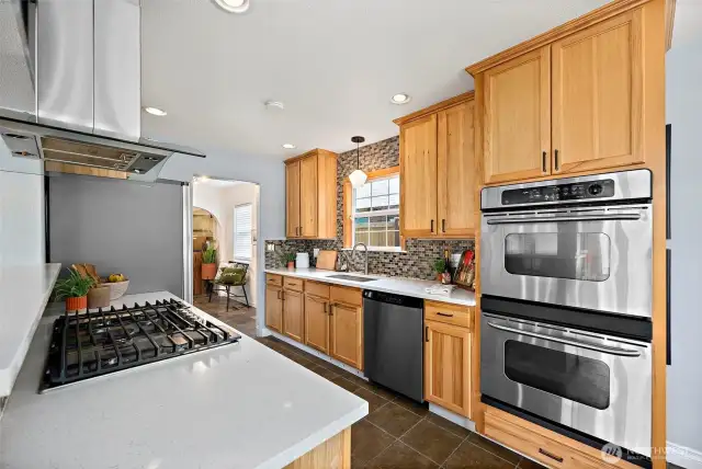 Galley kitchen with fridge, sink overlooking the backyard, gas stovetop, with double-ovens across from stove. View from kitchen entryway looking down kitchen to mudroom space.