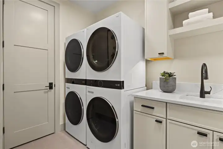 Laundry room with double stack LG front-loading washers and dryers. Upper and lower storage cabinets, slab quartz countertop, utility sink, open shelving with under counter lighting, and a linen closet.