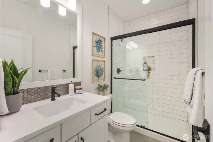 Lower Level Bathroom  This lower level main bathroom features a clean, modern design with quartz countertops, a sleek undermount sink, and matte black fixtures for contrast. A tiled shower with glass enclosure and built-in niche adds both function and architectural simplicity, while durable flooring supports everyday use.    Located in Bothell’s Canyon Park area, this full bath enhances the versatility of the lower level, allowing the adjacent den to function comfortably as a guest suite or additional bedroom. The thoughtful layout reflects the practicality and finish quality expected in modern new construction within the Northshore School District.    These photos are from a past Robinson Plan at a different community, so overall plan, colors, building orientation, finishes, home outlook, and other details can and WILL vary. All photos, renderings, site maps, and floorplans are for visualization purposes only.