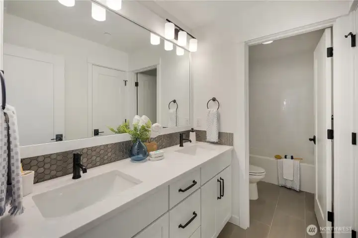 Upper Level Secondary Bathroom  This upper level secondary bathroom features a dual-sink vanity with quartz countertops, matte black fixtures, and a full-width mirror that enhances light and scale. Clean cabinetry lines and durable tile flooring create a streamlined, low-maintenance design suited for everyday use.    A separate enclosed tub and toilet area provides added privacy and functionality, supporting shared use with ease. Located in Bothell’s Canyon Park neighborhood, this well-appointed bath reflects the thoughtful planning associated with modern new construction homes served by the Northshore School District.    These photos are from a past Robinson Plan at a different community, so overall plan, colors, building orientation, finishes, home outlook, and other details can and WILL vary. All photos, renderings, site maps, and floorplans are for visualization purposes only.