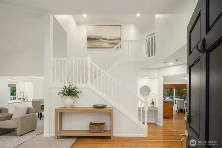 The entry way is impressive with double height vaulted ceilings.  The circular floorplan of the home is immediately obvious with formal living and dining to the left, and the kitchen, family room and den at the back of the house to the right. Console and artwork on the stairs have been added with AI