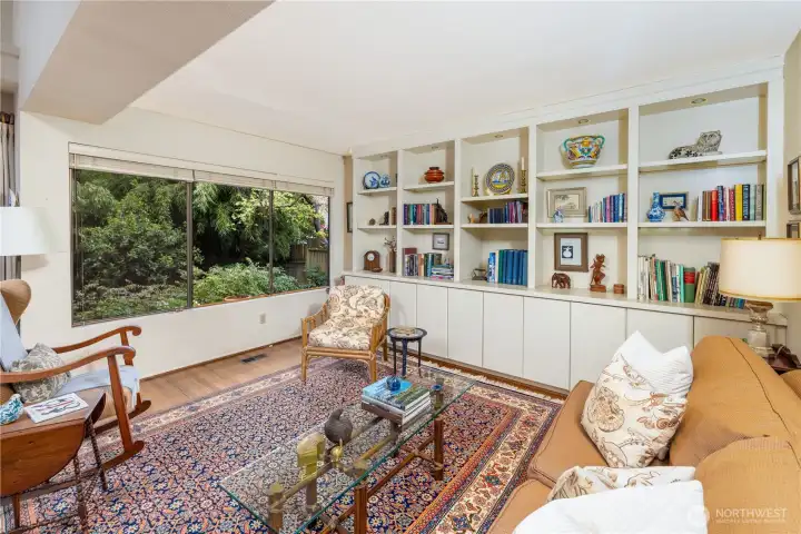This cozy sitting area is anchored by a wall of custom built-in bookcases, offering both charm and abundant storage. A large picture window frames serene garden views, filling the space with natural light and creating a peaceful spot for reading or relaxing.