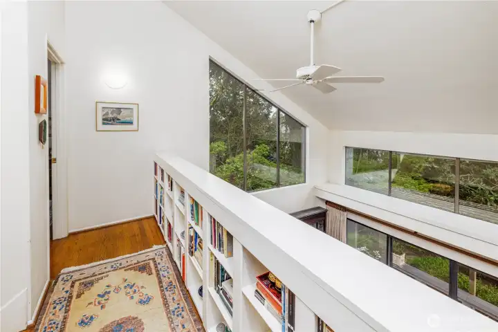 The second floor hallway features custom built-in bookshelves and overlooks the dramatic living area below, enhancing the home’s open, architectural design. Walls of windows bring in natural light and leafy views, creating a bright and visually striking transition between the bedrooms.