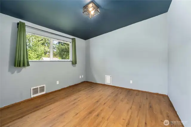 This generously sized bedroom features stylish laminate flooring, a bold accent ceiling, and a large window that fills the space with natural light and views of the lush outdoors. A cozy and quiet retreat at the end of the day.