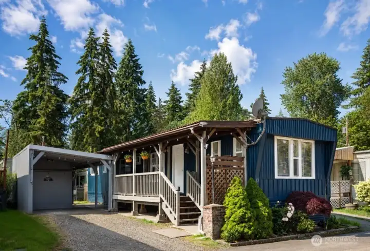 Front exterior of the single-wide home, featuring a covered entry, mature trees, and detached two small storage units.