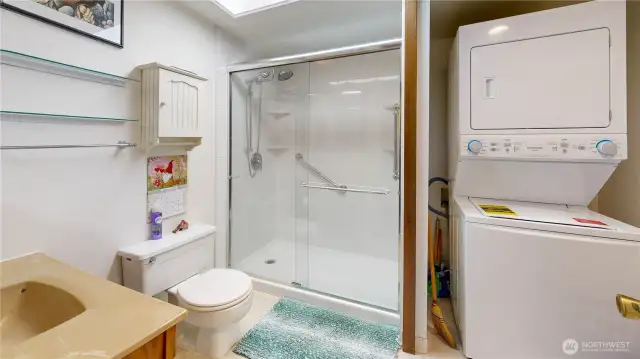 Well-appointed primary bath featuring a spacious step-in shower with glass enclosure and grab bars for added comfort. Includes built-in shelving, overhead skylight for natural light, and convenient in-unit laundry with a stacked washer and dryer.