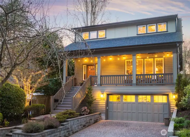 A warm twilight shot captures the home's covered front porch, paver driveway, attached garage, and thoughtfully landscaped entry.
