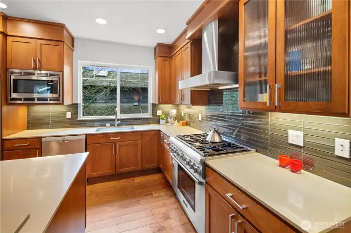 A close-up of the kitchen's cooking zone, featuring a professional-grade stainless range beneath a wall-mounted stainless hood, surrounded by a horizontal tile backsplash in a warm green-gray tone. A window above the sink brings in natural light.