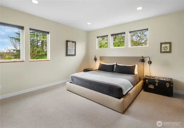 The primary bedroom occupies the upper level of the home and features plush carpet, warm cream-toned walls, and three windows providing treetop views. Wall-mounted sconces flank the bed wall.