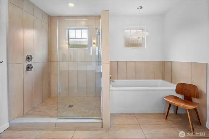 The shower and soaking tub area of the primary bathroom is clad floor-to-ceiling in large-format warm sand-tone tile. A glass-enclosed walk-in shower and a drop-in soaking tub occupies the other side.