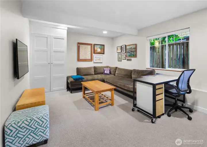 A room on the lower level offers flexible use, with a wall-mounted TV location, a gallery wall, and a window providing natural light. Two panel doors provide closet or storage access.