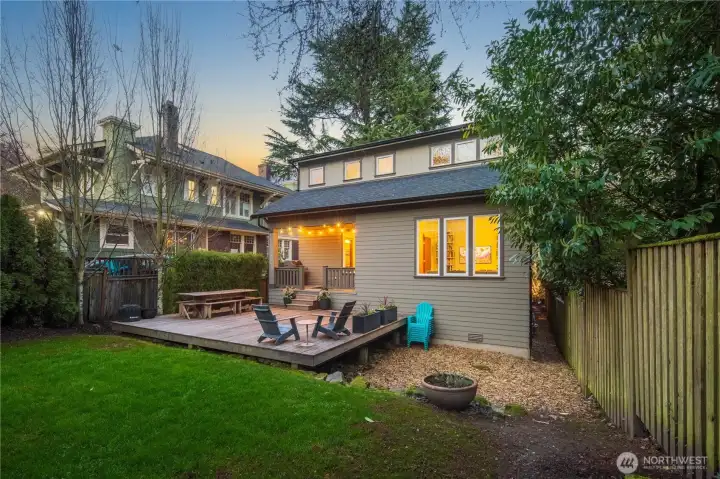 The rear yard features a wood deck adjacent to a fenced lawn area. String lights overhead and the warm glow of interior lighting through the back windows create an inviting outdoor atmosphere.