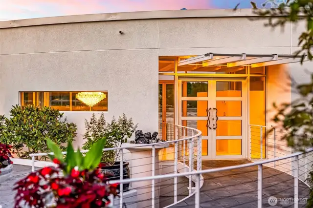 A dramatic entry sets the stage — the curved approach and glass double doors hint at the light-filled elegance within, as a sparkling chandelier glows like a beacon of luxury