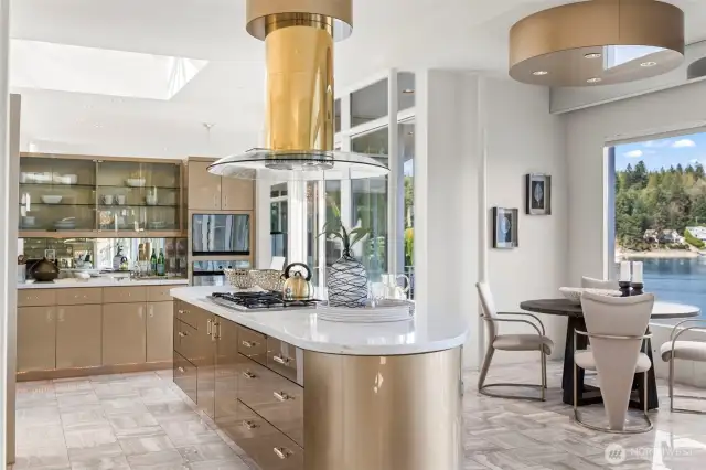 This designer Gig Harbor waterfront kitchen pairs sleek contemporary lines with warm, golden tones for a truly luxurious feel. A curved center island with marble countertop anchors the space, complemented by a sculptural range hood and high-gloss cabinetry. Walls of glass invite in natural light and frame serene views of the harbor, while an adjacent breakfast nook offers the perfect spot for morning coffee with the water as your backdrop. Ideal for both entertaining and everyday living, this kitchen blends style, functionality, and an unmistakable sense of place.