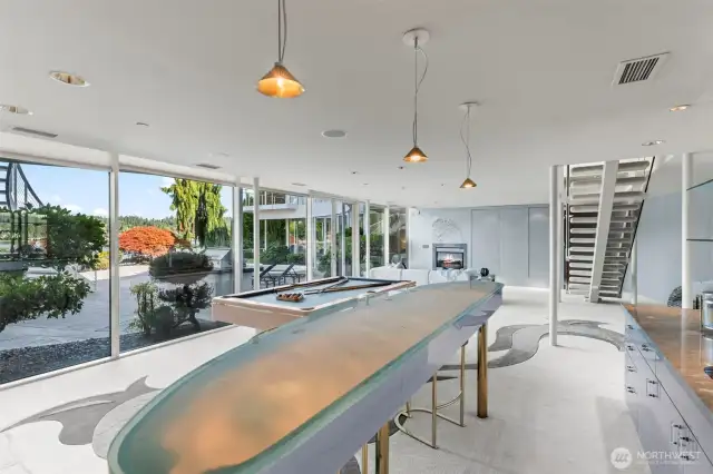 The recreation level features a sleek glass-topped bar cocktail table, a pool table, and walls of windows that open to the patio and waterfront.