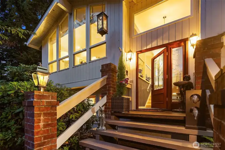 A warm welcome awaits! The inviting front entry showcases glowing lanterns, brick accents, and a stunning glass-paneled door that sets the tone for what’s inside. Elevated architecture and large picture windows capture the home’s signature mid-century charm while filling the interior with golden light. Every detail of this entry—from the stately pillars to the lush greenery—creates a memorable first impression that feels both elegant and inviting. ?????
