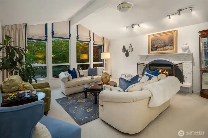 Vaulted ceilings, expansive picture windows, and soft natural light create a bright and welcoming gathering space. The cozy fireplace with white brick surround adds timeless character and warmth—perfect for relaxing evenings or entertaining in style.